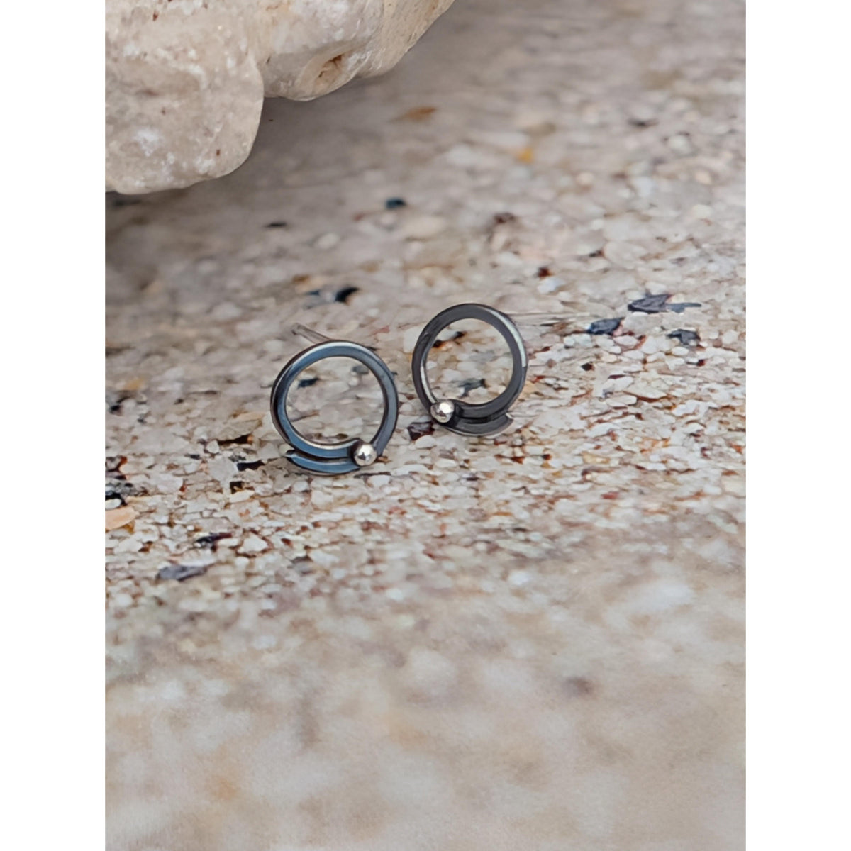 SA Ea328 wave hoop studs with silver granulation by Sandra Austin jewellery, available at Padstow Gallery, Cornwall. A pair of dark metallic ring earrings with small bead details, resting on a textured sandy surface.