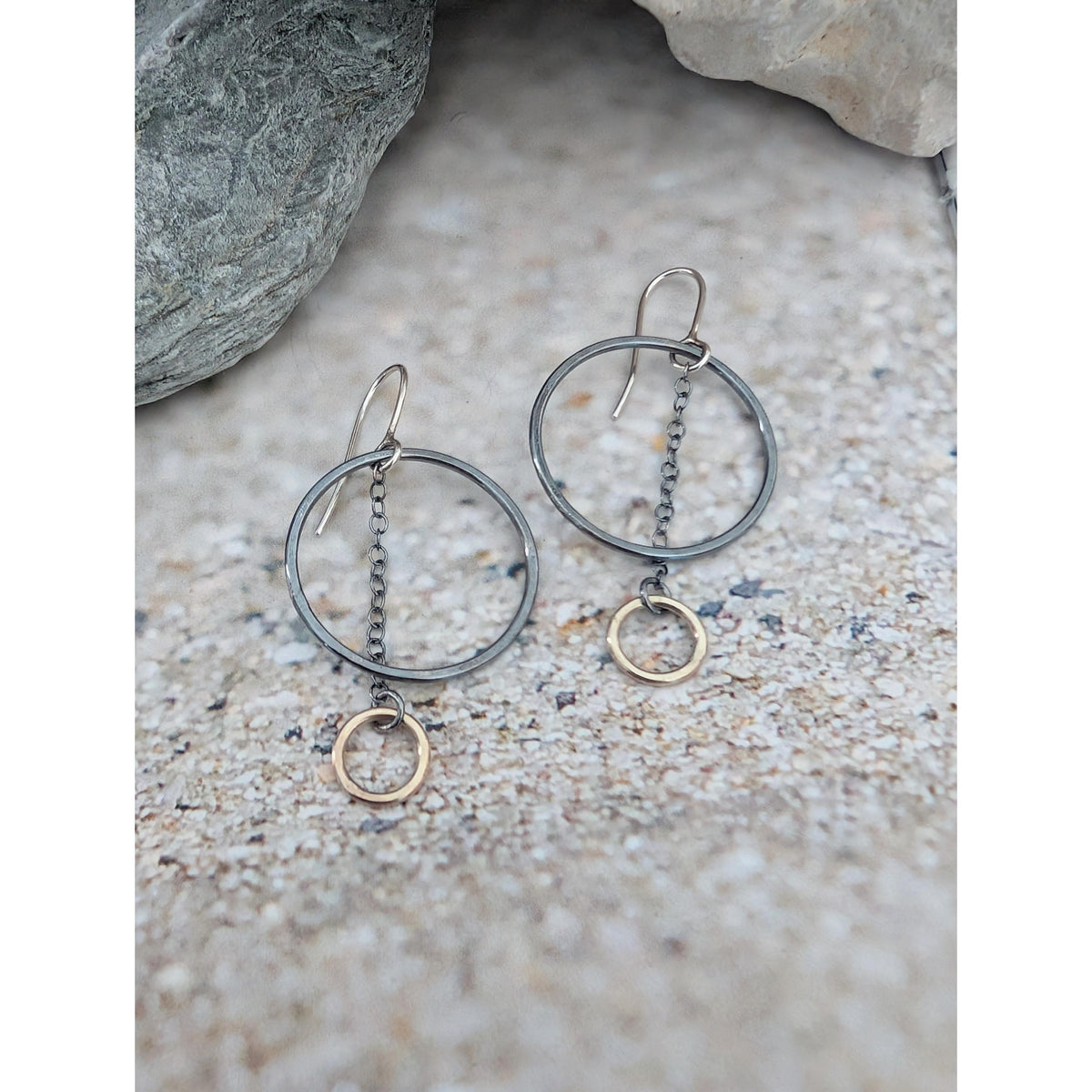 A pair of dangling earrings with oxidized silver-tone double hoop rings and small gold-tone circular charms, displayed on a textured stone surface.