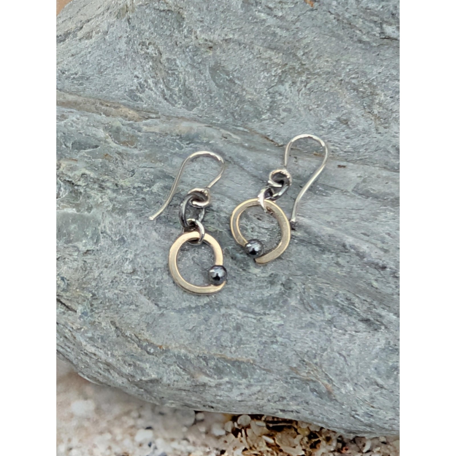 A pair of handmade dangle earrings with one matte gold-toned circle and one oxidized silver-toned circle, each with a small dark bead, resting on a textured gray stone.