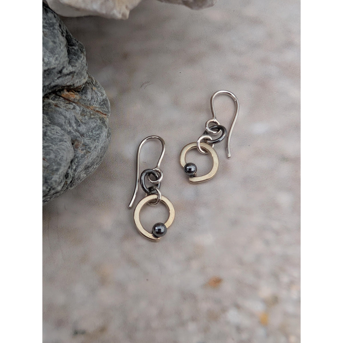 A pair of handmade dangle earrings with silver fishhook wires, small dark beads, and gold-toned circular elements, displayed on a textured stone surface next to a gray rock.