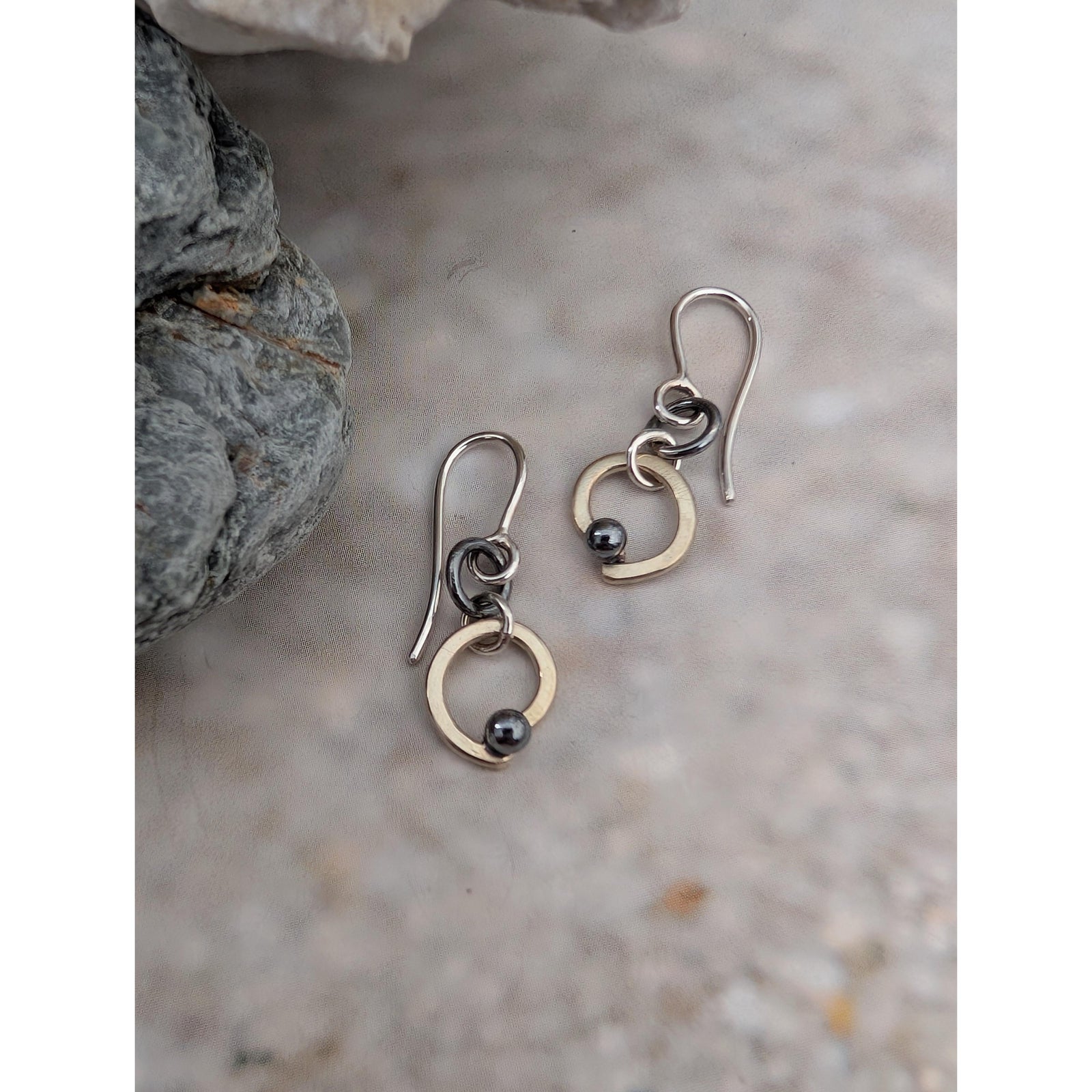 A pair of handmade dangle earrings with silver fishhook wires, small dark beads, and gold-toned circular elements, displayed on a textured stone surface next to a gray rock.
